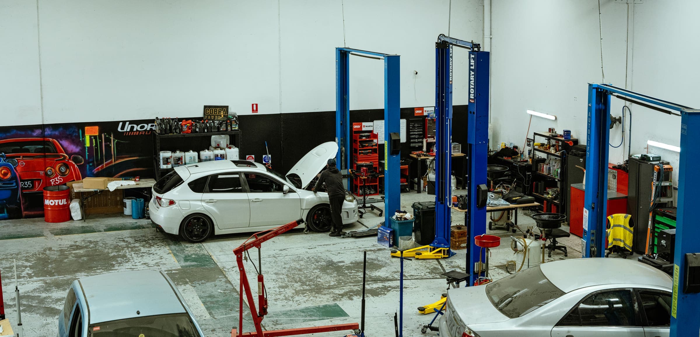 Overhead view across the Unorthodox Automotive workshop floor, showing both hoists and vehicles in for service